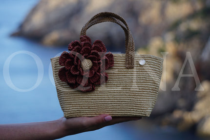 Sac en raphia beige avec anse marron et fleur déco rouge tenue à la main en bord de mer