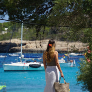 Femme debout de dos avec jupe blanche et sac en paille face à la mer turquoise et voilier